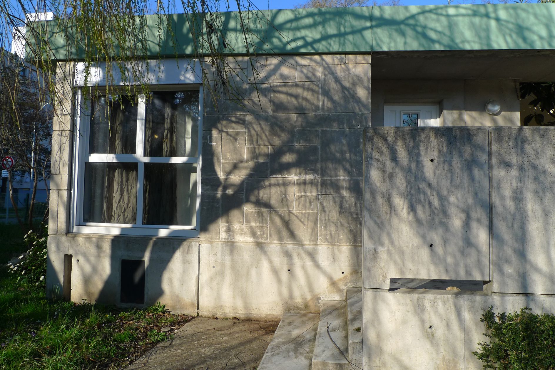 Façade de l’entrée. Accès à la porte par trois marches donnant sur un espace semi-couvert, délimité par un muret haut et un pilier métallique.