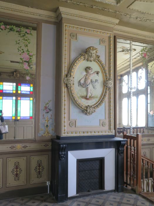 Salle des fêtes du 1er étage. Mur est. Cheminée en marbre noir, à consoles sculptées surmontées d’un angelot distribuant des fleurs dans une cadre ovale sommé par un faune grimaçant.