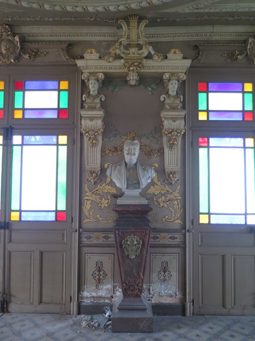 Salle des fêtes du 1er étage. Mur ouest. Panneau central. L’industriel Michel Sabatier est représenté en buste (par Louis Oury) sur un socle en marbre rouge orné d’un lion portant la date de 1899, sous une composition de 2 têtes cariatides portent un entablement couronné d’une lyre, symbole de son goût pour la musique.