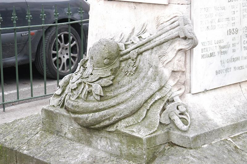 Vue de la base du piédestal, face antérieure, le casque, un rameau d'olivier, une croix de légion d'honneur posés sur un drapeau replié ; deux pommeaux de sabres sortent sous le drapeau.