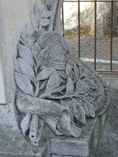 Vue de la base du piédestal, face antérieure, le casque, un rameau d'olivier, une croix de légion d'honneur posés sur un drapeau replié ; deux pommeaux de sabres sortent sous le drapeau.