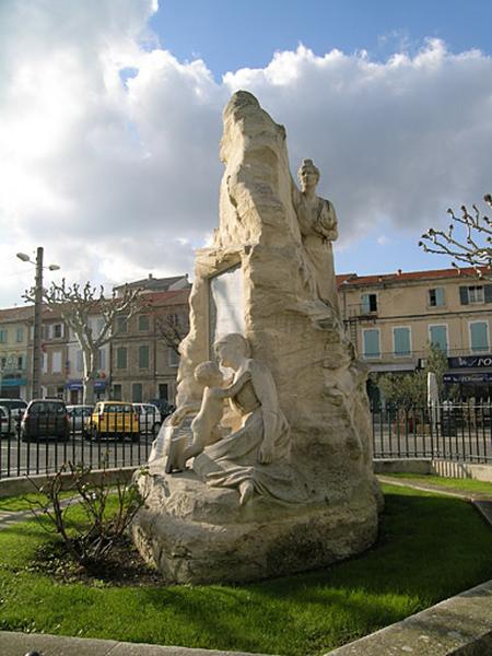 Vue générale de profil, le monument en forme de rocher émerge d'un terre-plein circonscrit par trois gradins en pierre dessinant un octogone ; il est entouré d'une grille.