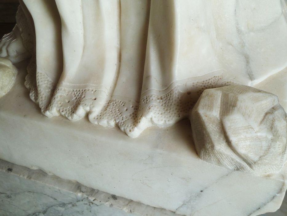 Statue de Saint Étienne : détail de la dentelle qui orne la partie inférieure de l’aube et des pierres posées à ses pieds sur le côté droit.